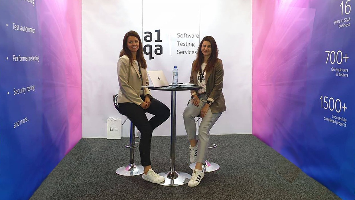Two staff at a1qa Software Testing Services exhibition booth with laptop and company branding