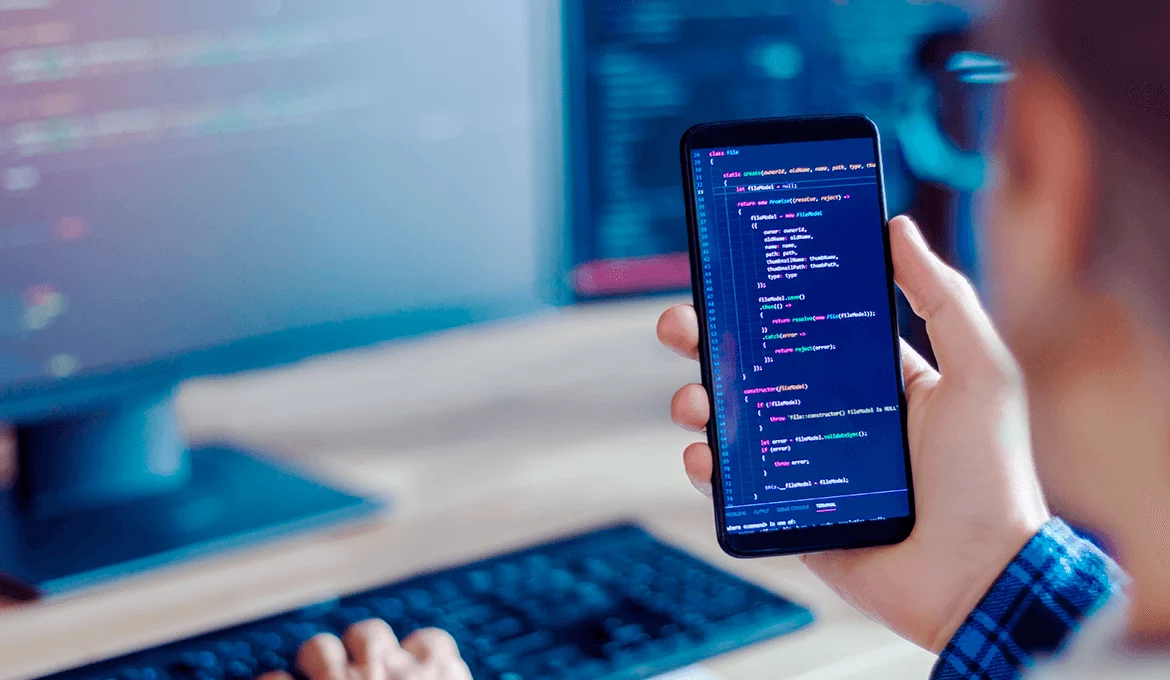 Developer reviewing code on smartphone beside desktop monitor, illustrating web service testing