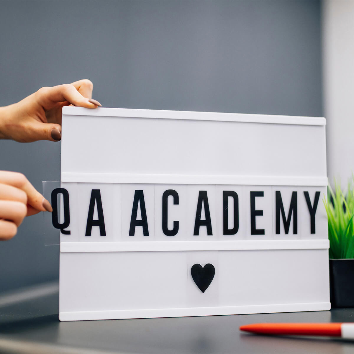 Hands placing letters on a lightbox sign reading QA ACADEMY with a heart icon
