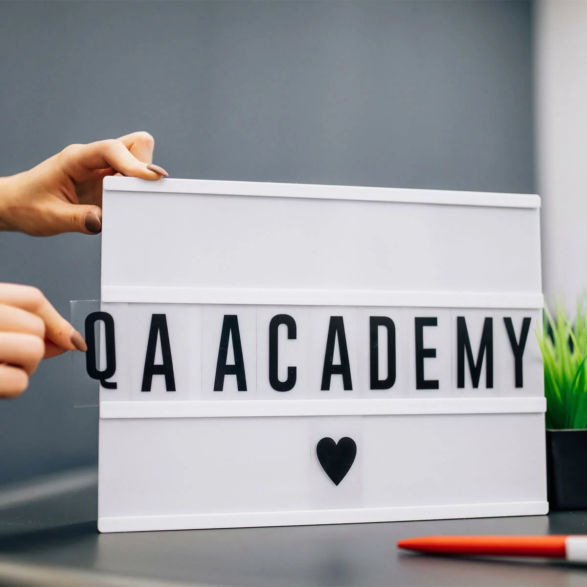 Hands placing letters on a lightbox sign reading QA ACADEMY with a heart icon
