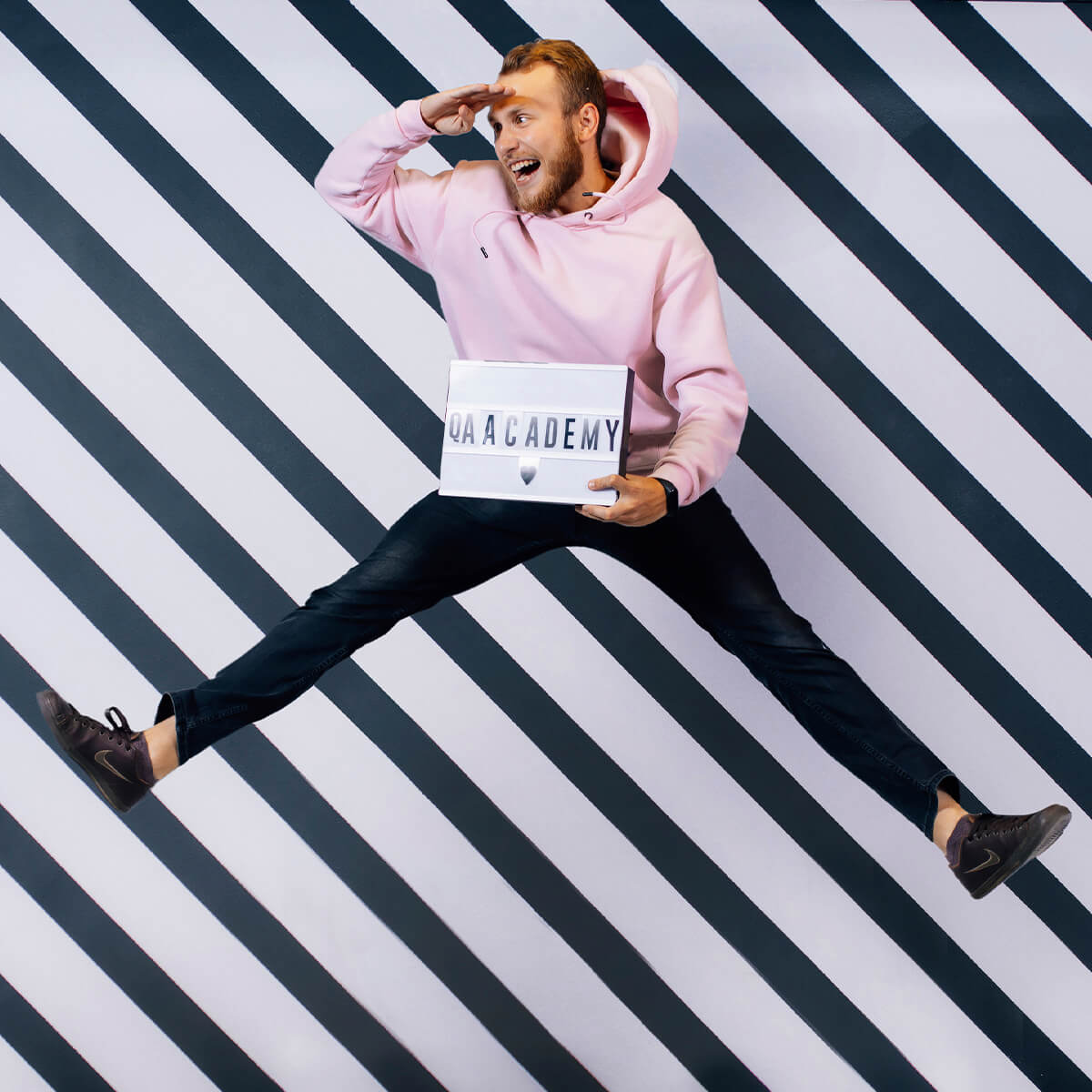Man in pink hoodie jumping against striped wall, holding QAACADEMY lightbox and looking ahead