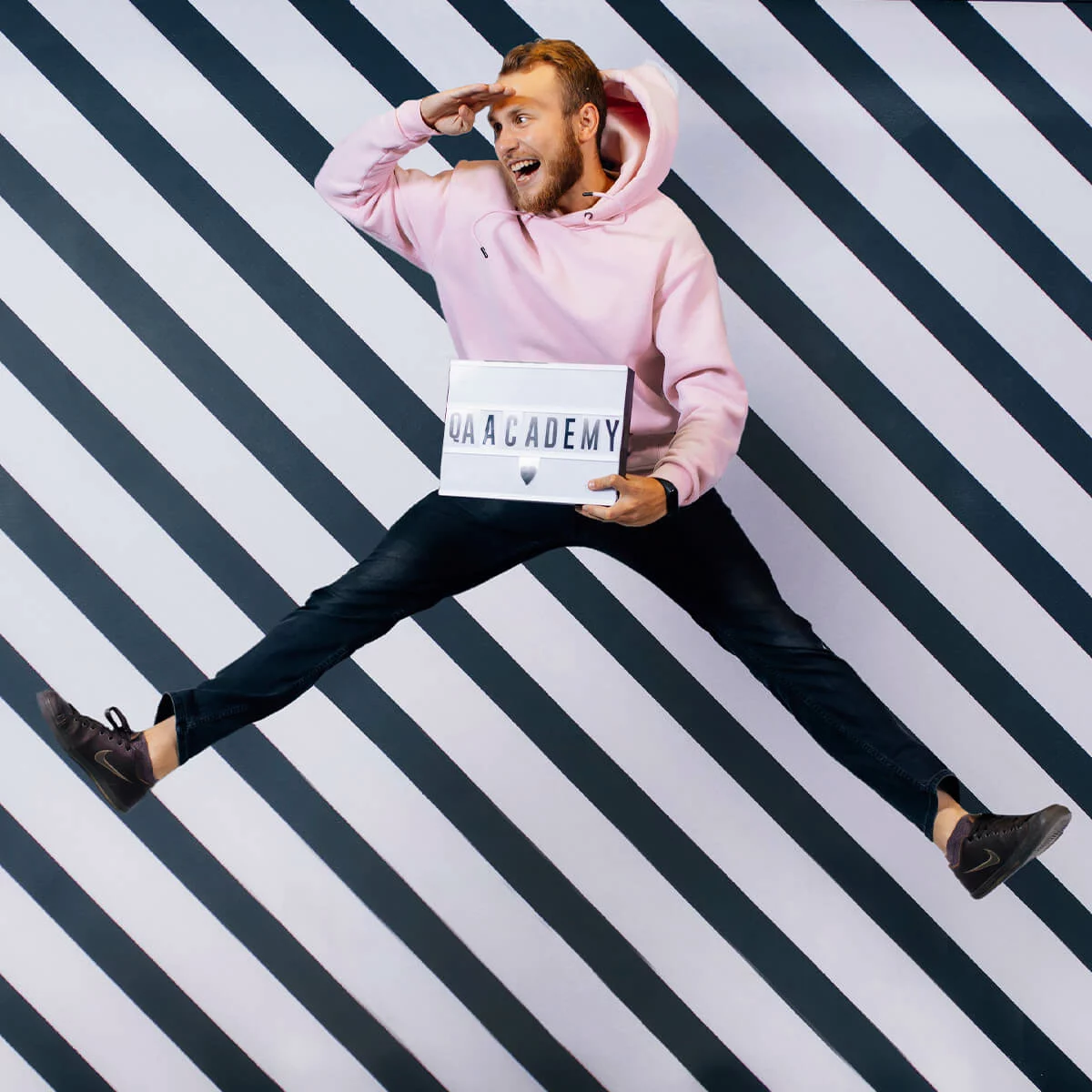 Man in pink hoodie jumping against striped wall, holding QAACADEMY lightbox and looking ahead