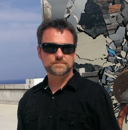 Man in sunglasses and black shirt standing outdoors beside a fragmented mirrored wall