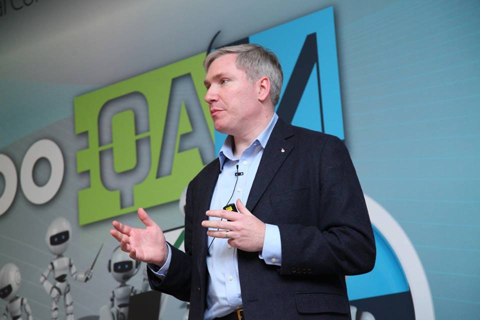 Matthew Heusser speaking at an event in front of the a1qa logo and robots