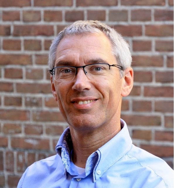 Portrait of a smiling man wearing glasses and a light blue shirt against brick wall