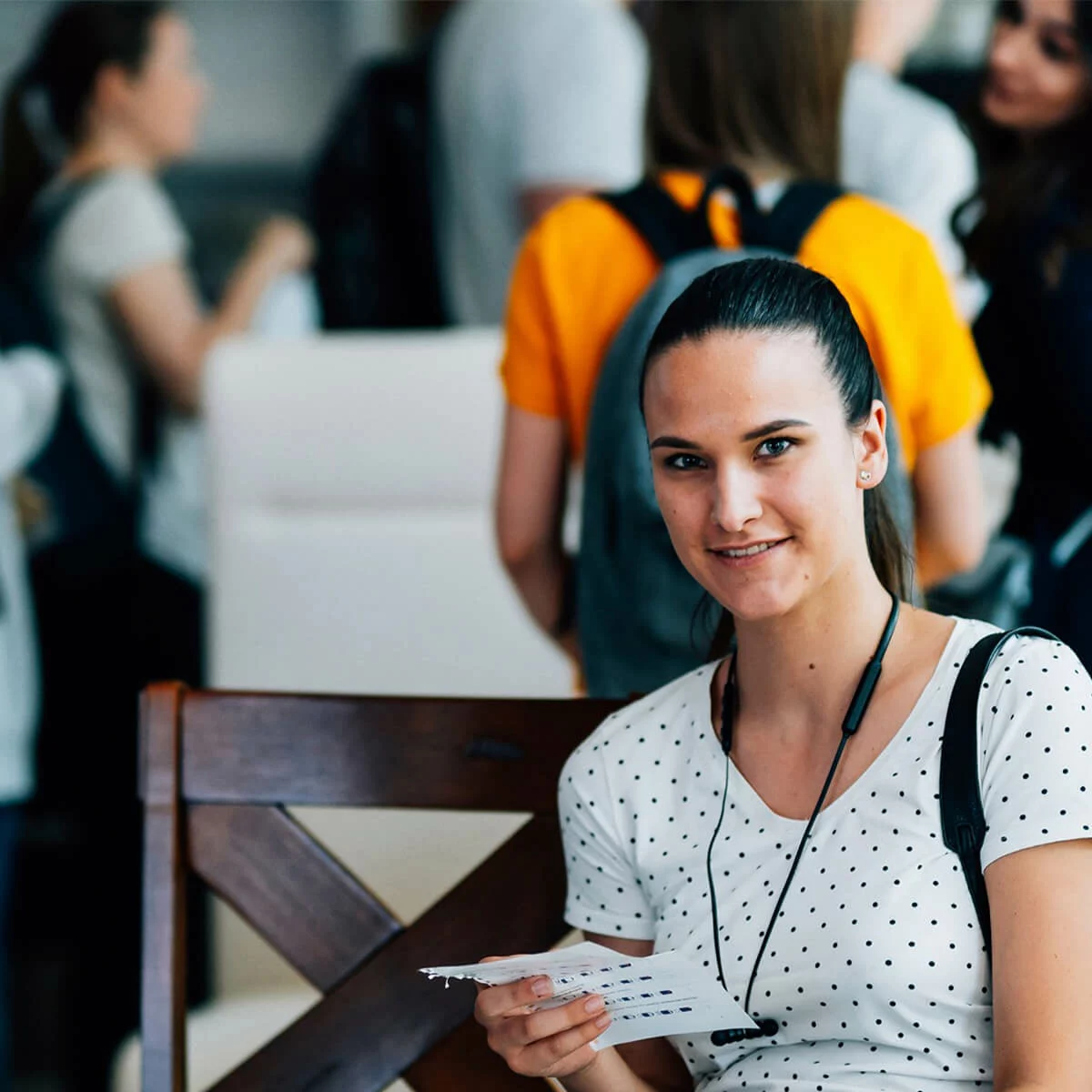 Young woman holding papers and wearing earphones in a busy training or academy setting
